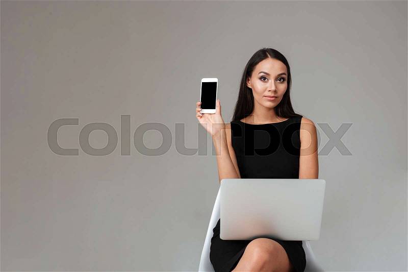 Young brunette woman in black dress sitting with laptop and showing blank screen mobile phone over gray background, stock photo
