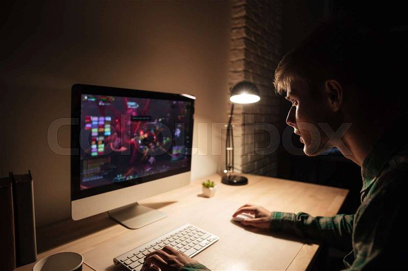 Handsome young man sitting and playing computer games in dark room, stock photo