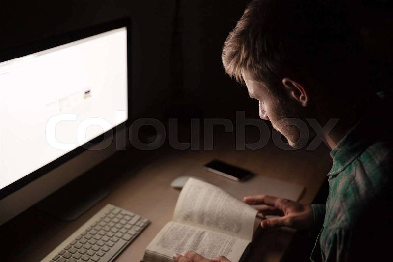 Serious young man reading book and using computer in the dak room, stock photo