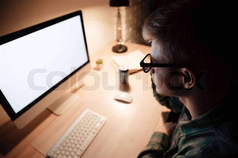 Serious young man in glasses working with black screen computer in the evening, stock photo