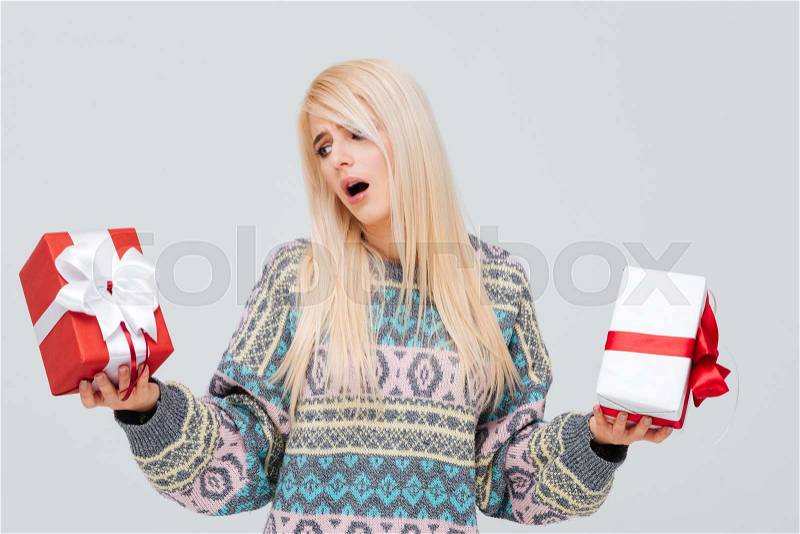 Frustrated blonde girl in sweater choosing gift boxes in her hands isolated on the gray background, stock photo