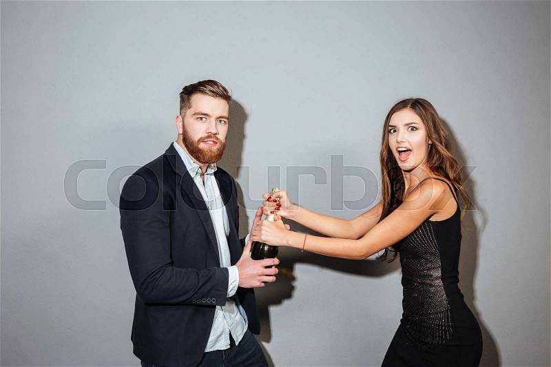 Young couple in formal wear fighting for champagne bottle over gray background, stock photo