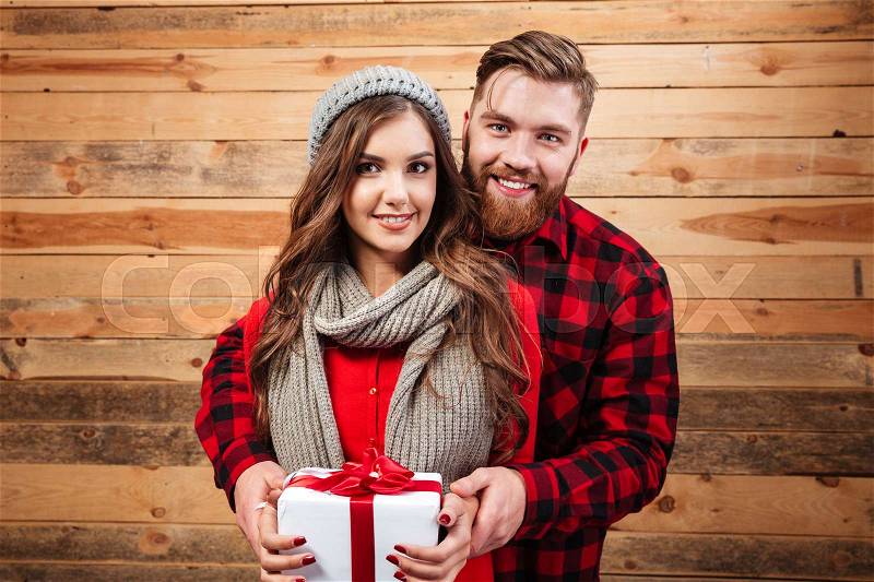 Happy young smiling couple in winter wear standing and holding gift box isolated on wooden background, stock photo