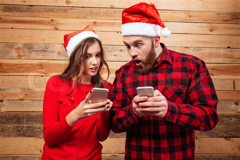 Friends in santa\'s hats with phones. looking at phones. shocked. wooden background, stock photo