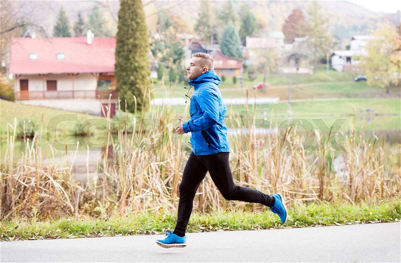 Young athlete in blue sports jacket at the lake running against colorful autumn nature, stock photo