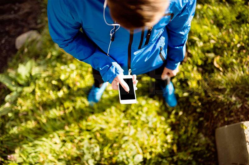 Unrecognizable runner with smart phone and earphones, listening music or using a fitness app. Using phone app for tracking weight loss progress, running goal or summary of his run. Outside in sunny autumn nature, stock photo