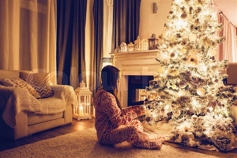Child in pajama decorating the Christmas tree in illuminated room some december night, winter weekends, cozy scene, stock photo