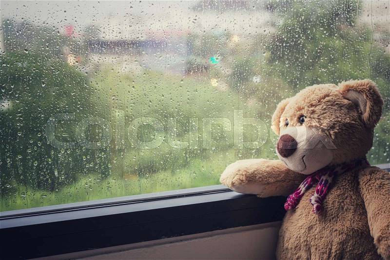 Brown teddy bear sitting beside the window while raining. Vintage filter effect, stock photo