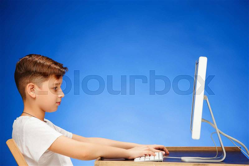School-age boy sitting in front of the monitor laptop at blue studio backgroubd, stock photo