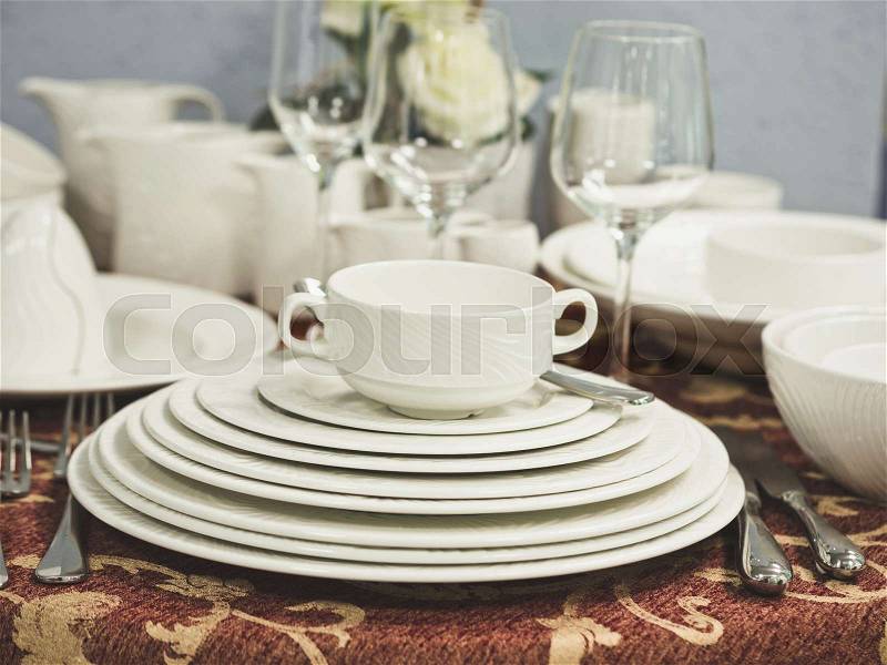Set of new dishes on table with tablecloth. Stack of white plates and wine glasses with flowers on restaurant table. Shallow DOF, stock photo