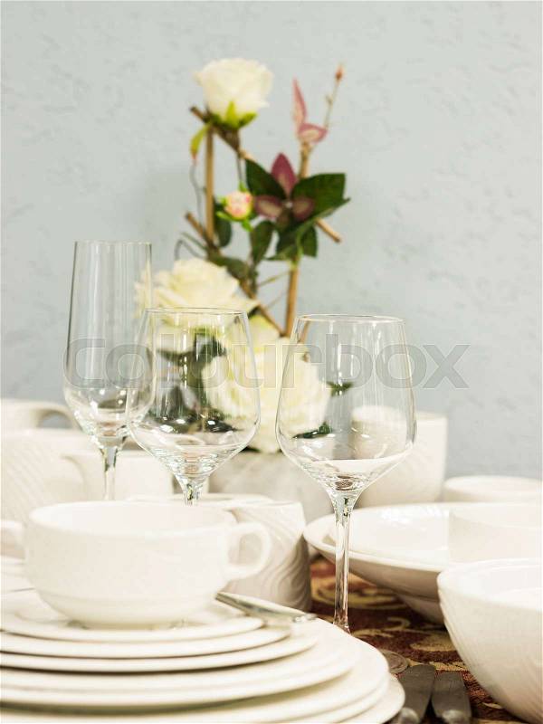 Set of new dishes on table with tablecloth. Stack of white plates and wine glasses with flowers on restaurant table. Vertical. Shallow DOF, stock photo
