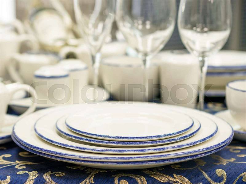 Set of new dishes on table with blue tablecloth. Stack of plates, saucer and wine glasses on restaurant table. Shallow DOF, stock photo