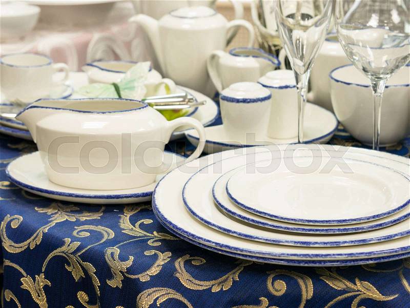 Set of new dishes on table with blue tablecloth. Stack of plates, saucer and wine glasses on restaurant table. Shallow DOF, stock photo