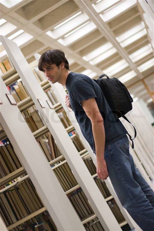 A young man reads a book at the library while standing in the aisles of book shelves, stock photo