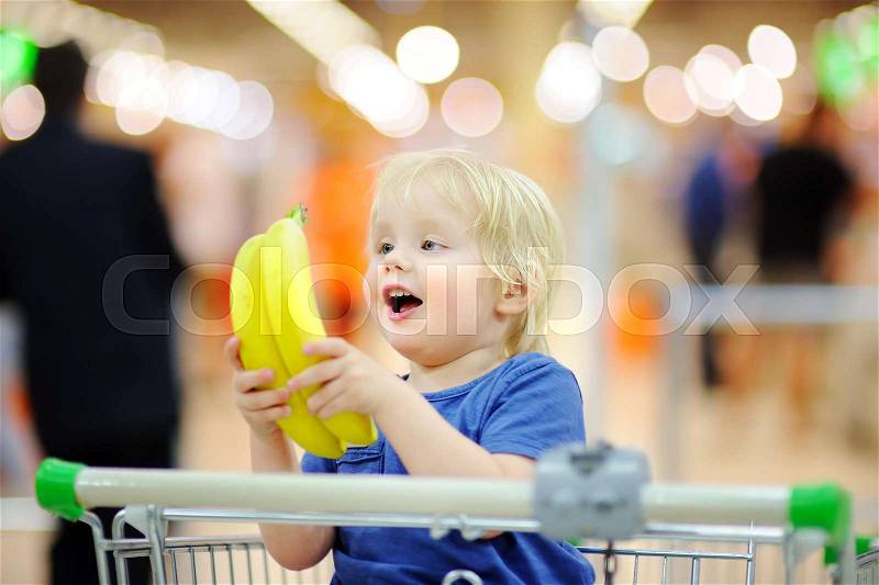 Cute toddler boy sitting in the shopping cart and holding bananas in a food store or a supermarket. Family shopping, stock photo