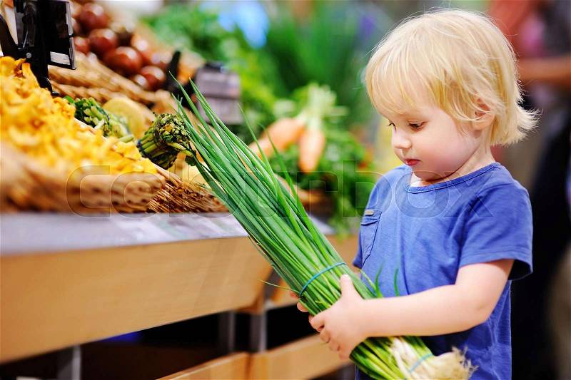 Cute toddler boy in a food store or a supermarket choosing fresh organic green onion. Healthy food for young family with kids, stock photo