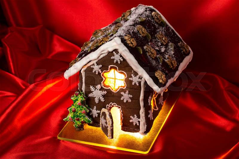 Christmas glazed gingerbread house with sweet pine and walnuts on housetop. Against red silk background, stock photo