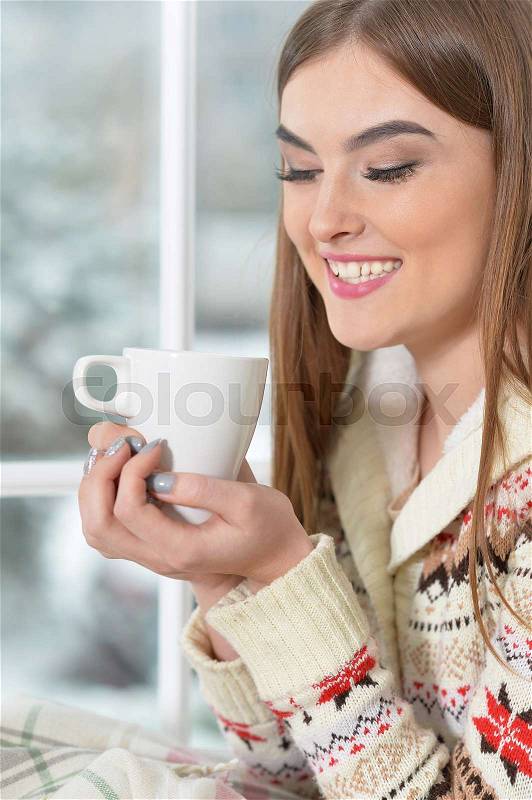 Beautiful girl in warm sweater drinking tea near the window, stock photo