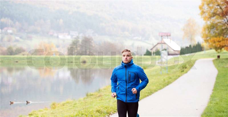 Young athlete in blue sports jacket at the lake running against colorful autumn nature, stock photo