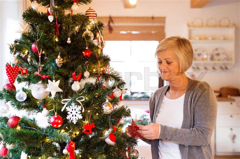 Beautiful senior woman decorating Christmas tree inside in her house, stock photo