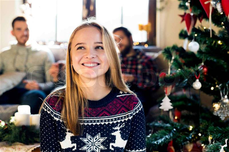 Young friends at decorated Christmas tree celebrating Christmas together. Teenage girl in sweater with nordic pattern smiling, stock photo