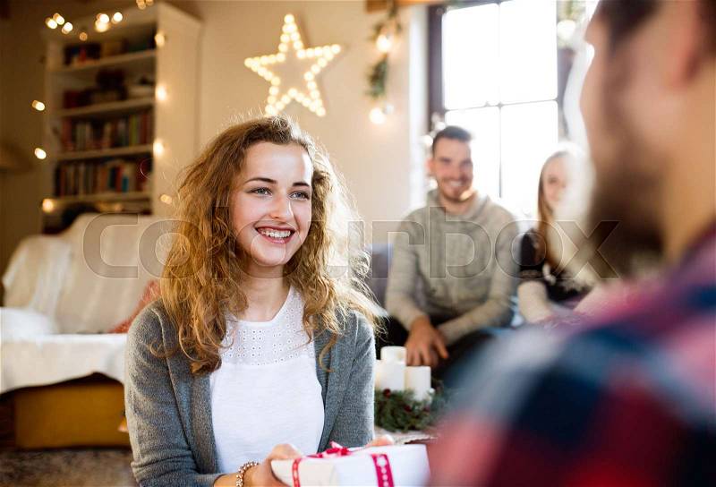 Young friends in decorated living room celebrating Christmas together exchanging gifts. Hipster man giving gift to beautiful young woman, stock photo