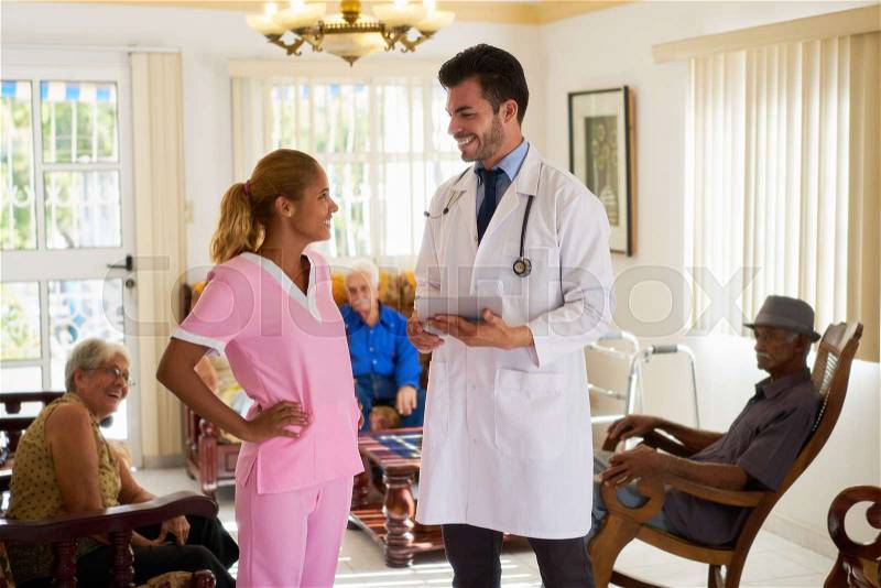 Doctor talking to young woman at work as nurse in hospital for seniors. The man holds a tablet pc. Some elderly people are sitting in background, stock photo