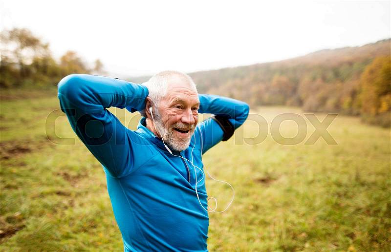 Senior runner in sunny autumn nature doing stretching. Man with earphones, listening music, armband on his arm. Rear view, stock photo