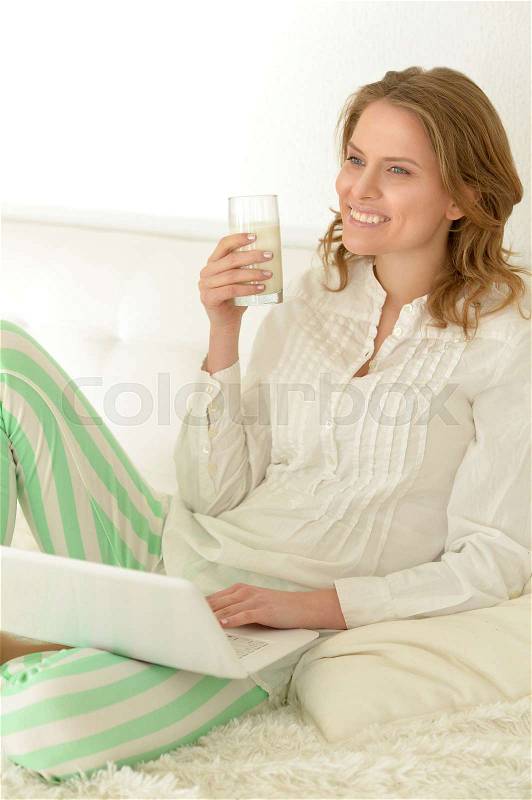 Beautiful young woman drinking milk and working on laptop on her bed at home, stock photo