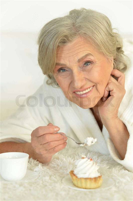 Portrait of seniour woman eating cake and drinking tea lying on the bed, stock photo