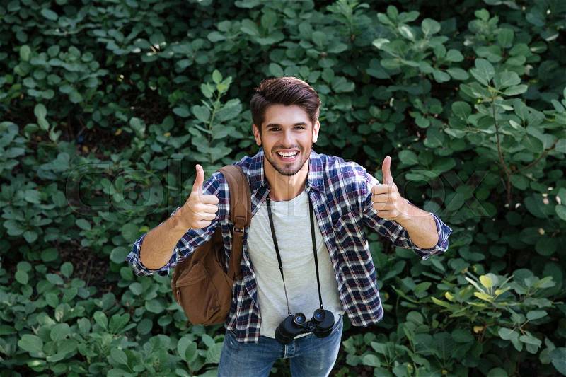 Happy man in shirt with binoculars and backpack in forest shows fingers up. looking at camera. top view, stock photo