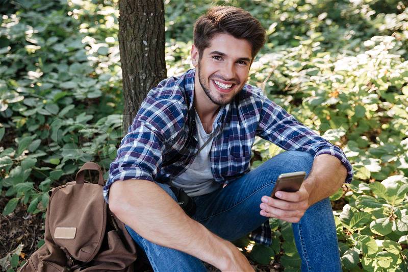 Happy man sitting in forest. top view. man with phone and backpack. near the tree. looking at camera, stock photo