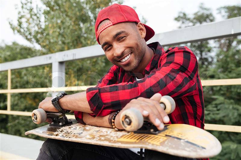 Photo of cheerful dark skinned man wearing cap holding skateboard. Against nature background. Looking at the camera, stock photo