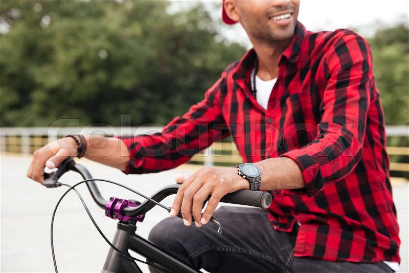 Cropped picture of happy dark skinned guy wearing cap sitting on his bicycle. Against nature background, stock photo