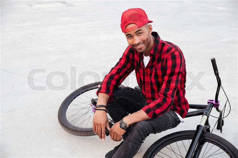Picture of happy dark skinned boy wearing cap sitting on his bicycle. Against nature background. Looking at the camera, stock photo