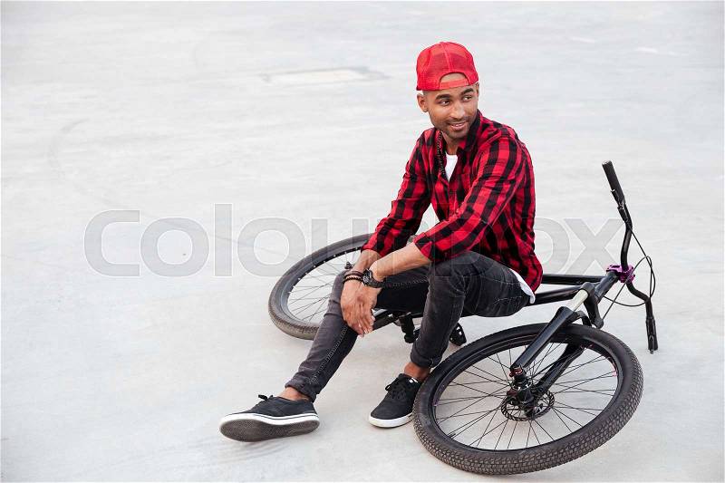 Picture of cheerful dark skinned guy wearing cap sitting on his bicycle. Against nature background, stock photo