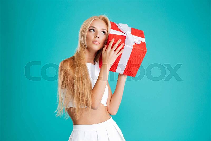 Portrait of a charming wondering woman holding gift box at her ear over blue background, stock photo