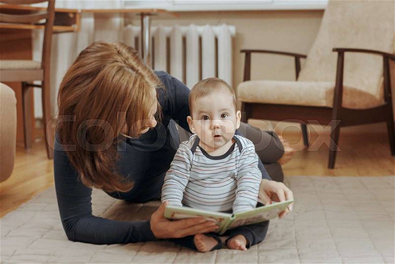 Young mother reading a story to her infant boy as they relax together on a carpet at home with the child following along in the book with a look of concentration, stock photo