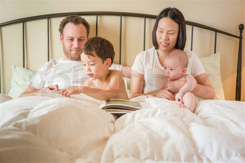 Young Mixed Race Chinese and Caucasian Baby Boys Reading a Book In Bed with Their Father and Mother, stock photo