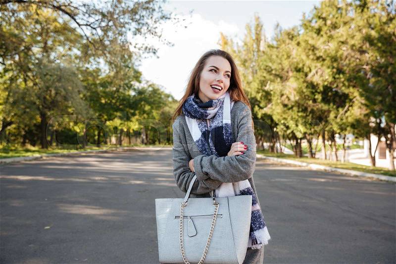 Portrait of happy young woman wearing scarf smiling against nature background and holding bag. Look aside, stock photo