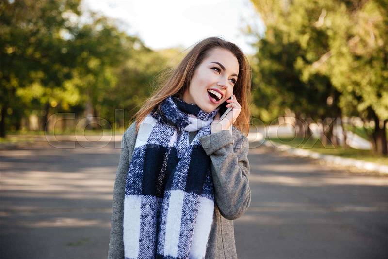 Photo of young happy woman wearing scarf talking on cellphone against nature background. Look aside, stock photo