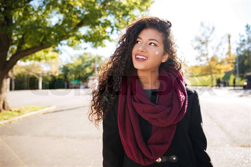 Photo of cheerful young dark skinned curly lady wearing scarf against nature background. Look aside, stock photo