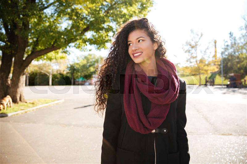 Portrait of happy young dark skinned curly woman wearing scarf smiling against nature background. Look aside, stock photo