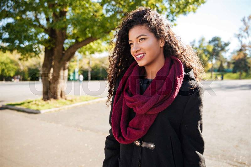 Portrait of cheerful young dark skinned curly woman wearing scarf against nature background. Look aside, stock photo