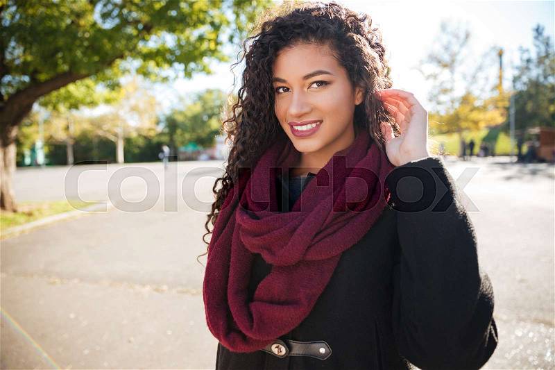 Photo of happy young dark skinned curly girl wearing scarf touching her hair against nature background. Look aside, stock photo