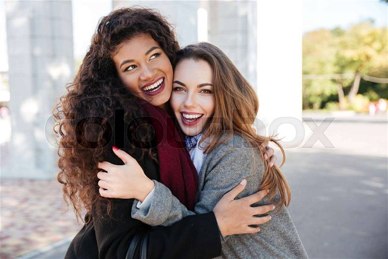 Picture of happy meeting of two friends hugging with street on background. Caucasian girl looking at camera. African lady look aside, stock photo