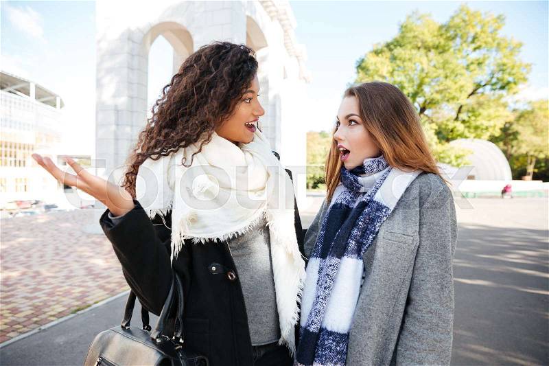 Two surprised young ladies looking at each other on street background with trees. Caucasian lady wearing scarf in a cage print. African young curly woman wearing white big scarf and holding bag, stock photo
