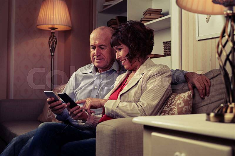 A mid shot of a pair communicating with one another, smiling and pointing at something funny on their mobiles. Elderly man and woman sitting in a room with an excellent modern interior, stock photo