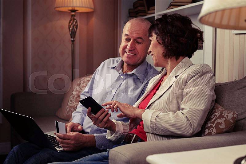 A mid shot of a smiling pair looking at something funny on the phone. People sitting on the settee with cute cushions, a laptop on man’s knees, stock photo