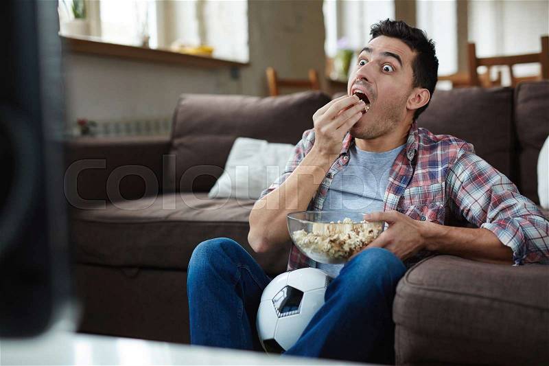 Astonished guy eating popcorn while watching curious tv program, stock photo